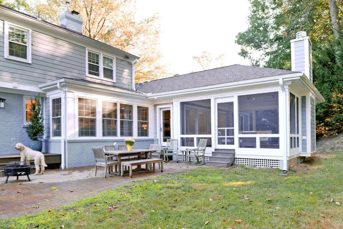 Backyard of a gray house with a screened porch, outdoor dining table, and a dog standing next to a grill.