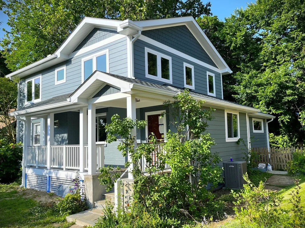 Two-story house painted blue with white trim, front porch, surrounded by green trees and plants.