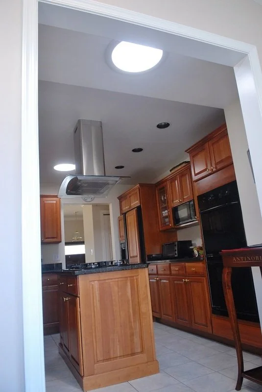 View of a kitchen with wooden cabinets, a black granite countertop, a stainless steel range hood, and black appliances. Part of a dining chair is visible in the foreground.