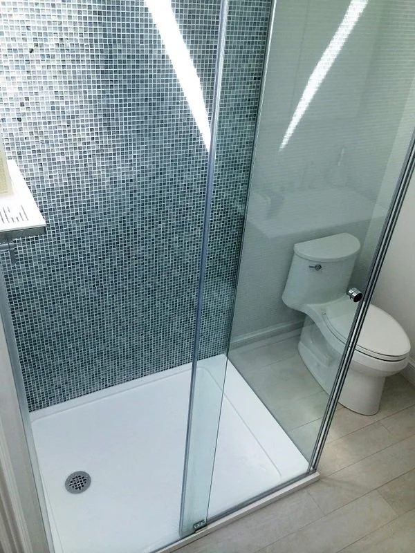 A bathroom with a glass shower enclosure and a white toilet. The shower has small blue mosaic tiles on the wall and a white shower base. The bathroom floor is light-colored wood.