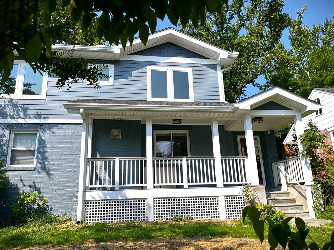 A two-story blue house with a front porch, white railing, steps leading to the door, surrounded by green trees and plants.