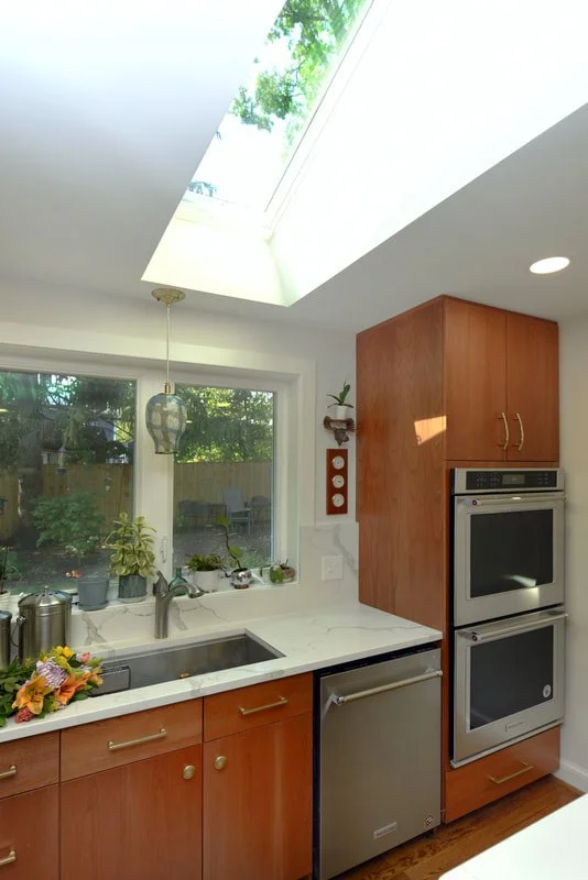 Modern kitchen with wooden cabinets, stainless steel oven and dishwasher, window above sink with plants, skylight in ceiling, and a hanging light fixture.