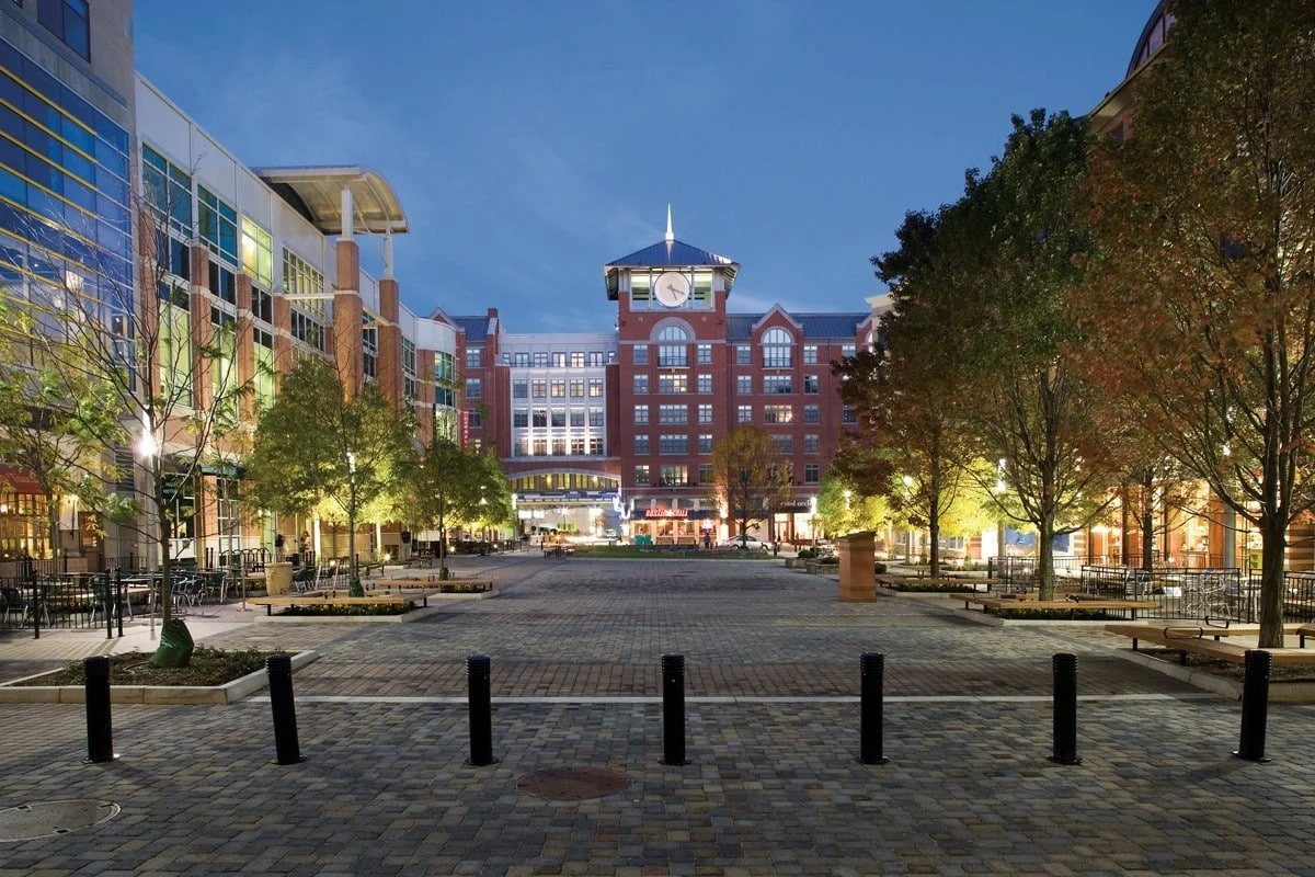 City square at dusk with trees, benches, and modern buildings, including a central clock tower.