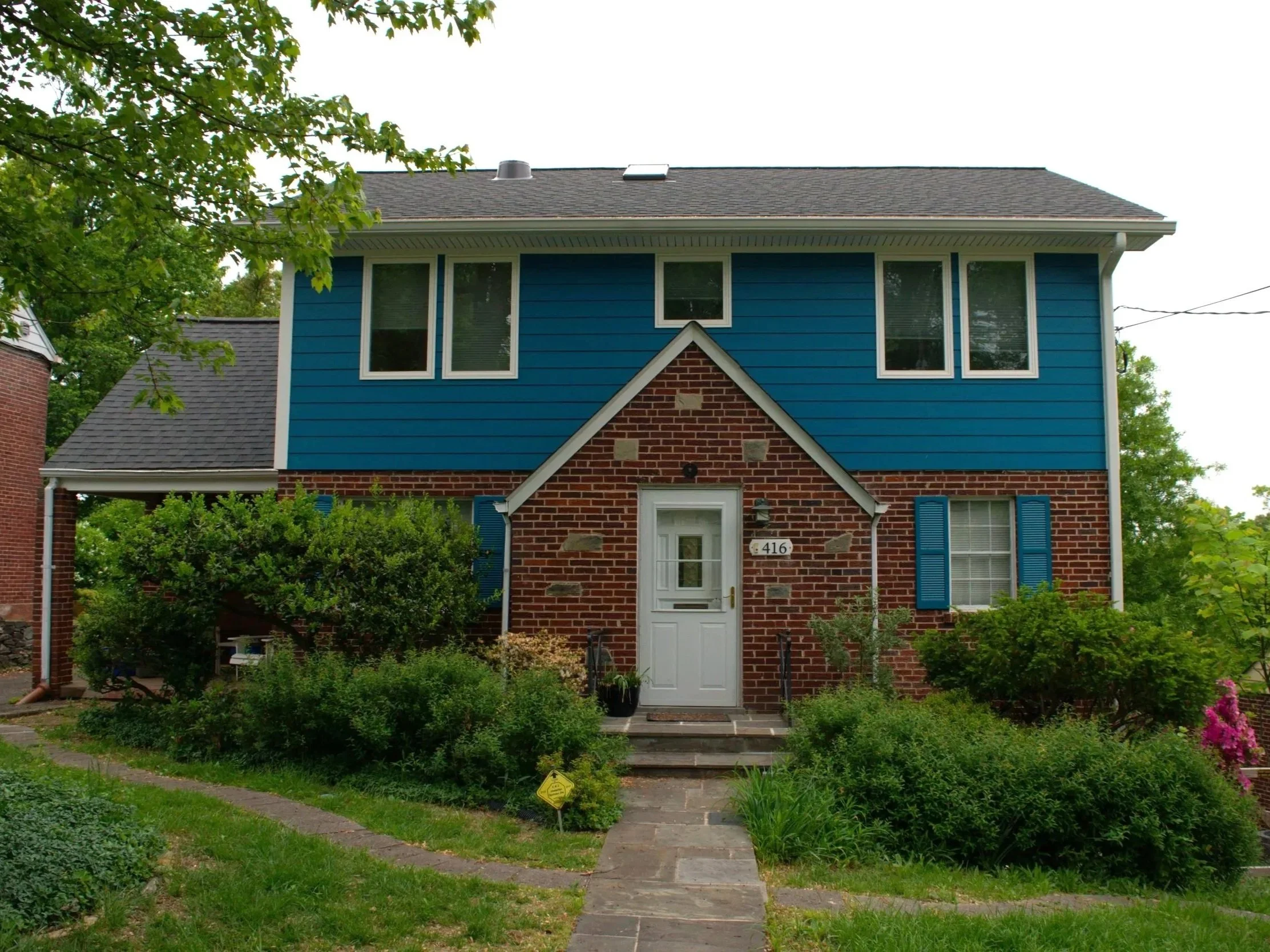 A house with a combination of brick and blue siding, multiple windows, a front door, and a garden with bushes, trees, and a walkway.