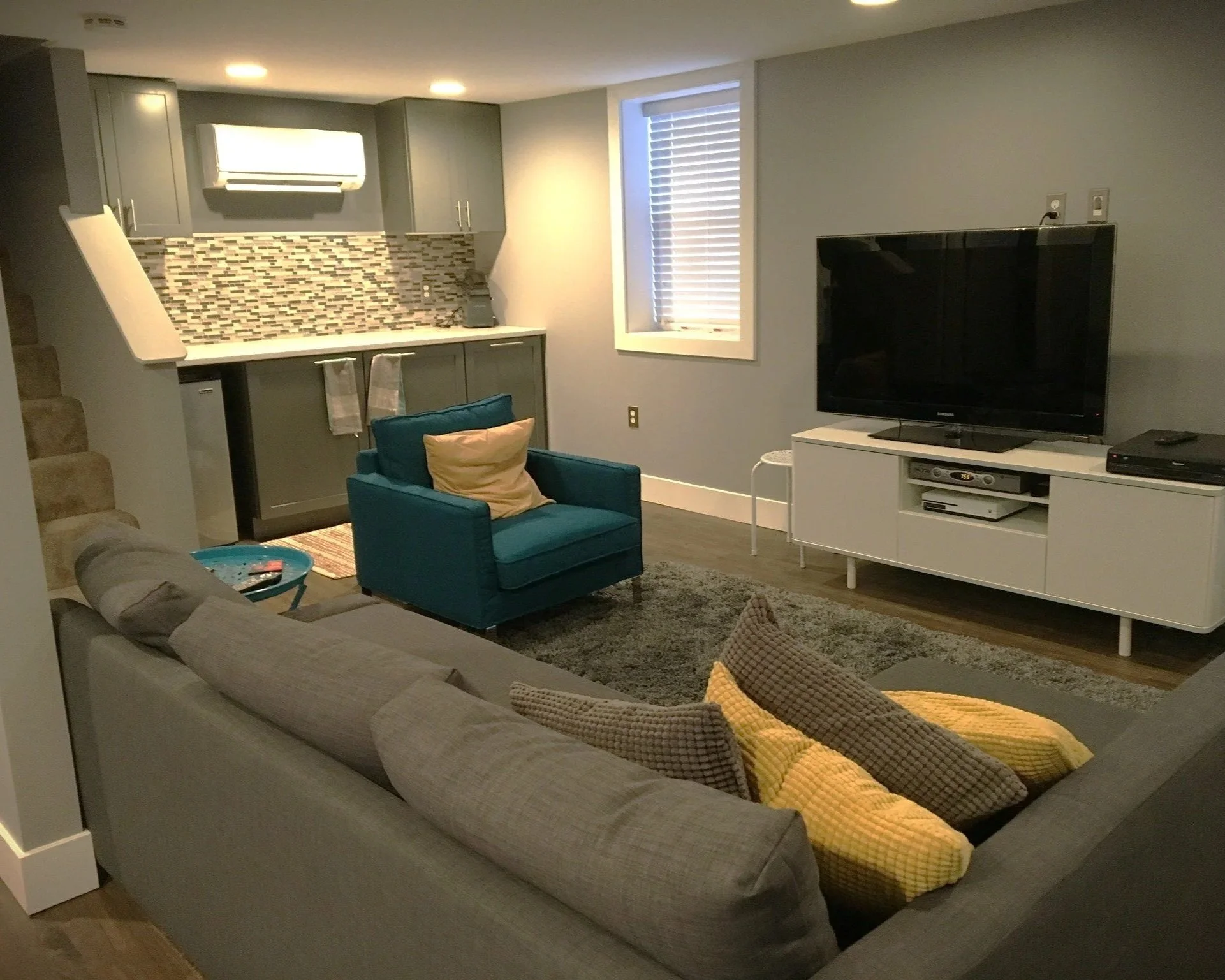Living room with a beige sofa, teal armchair, and a white TV stand with a flat screen TV. The room features a window with blinds, a gray area rug, and direct access to a small kitchen with gray cabinets, mosaic tile backsplash, and an air conditioning unit.