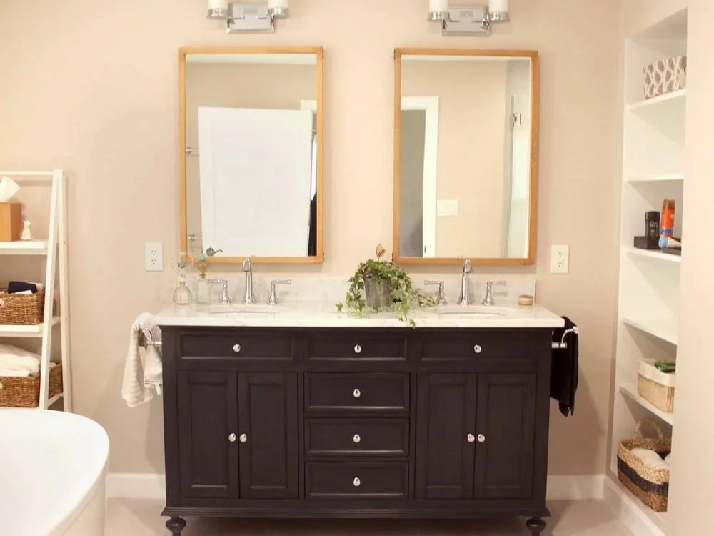 Bathroom vanity with two mirrors, a black cabinet, and a white countertop with two sinks, plants, and toiletries.