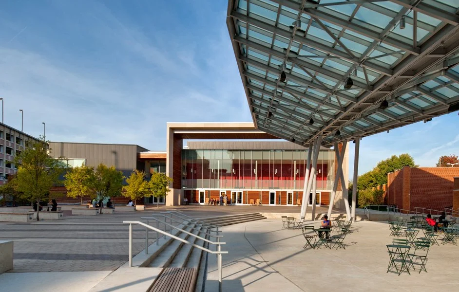 Modern outdoor plaza with a large glass and metal canopy, a building with glass windows, trees, and a few people sitting at outdoor tables under a clear sky.
