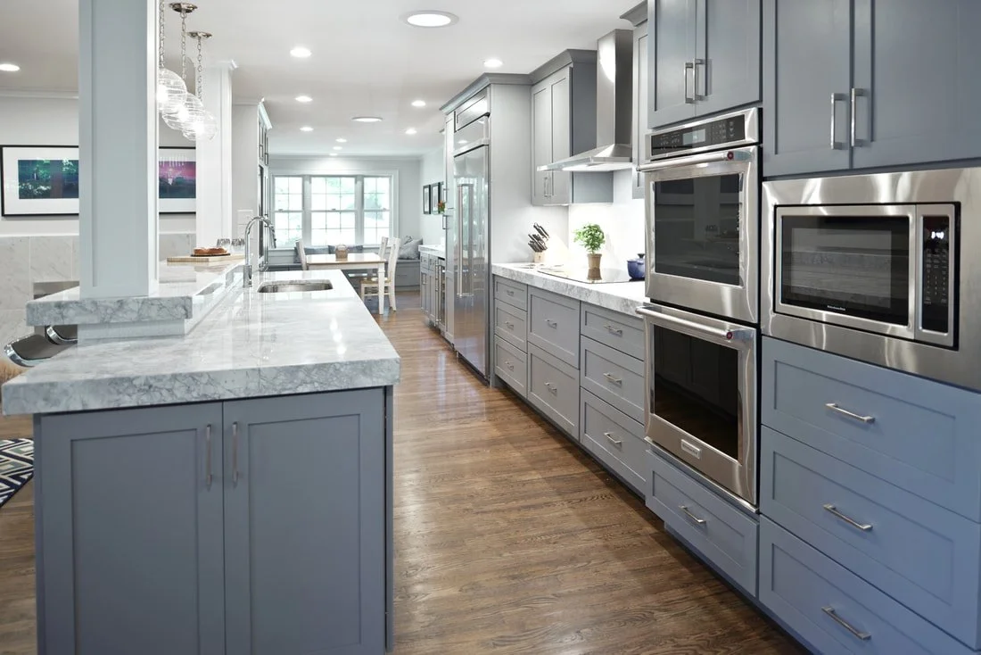 Modern kitchen with gray cabinets, stainless steel appliances, marble countertops, and hardwood flooring, with a dining area in the background.