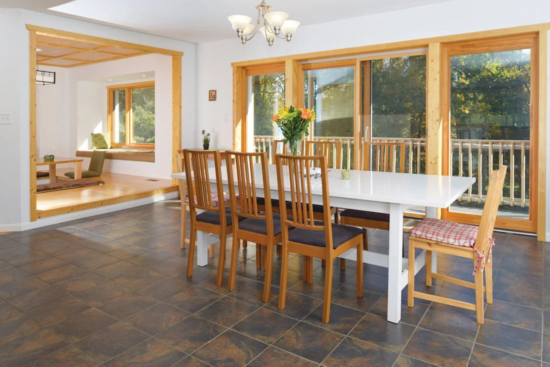 Dining room with large white table, six black-cushioned wooden chairs, flower vase, and sliding glass doors with wood framing leading to a balcony.