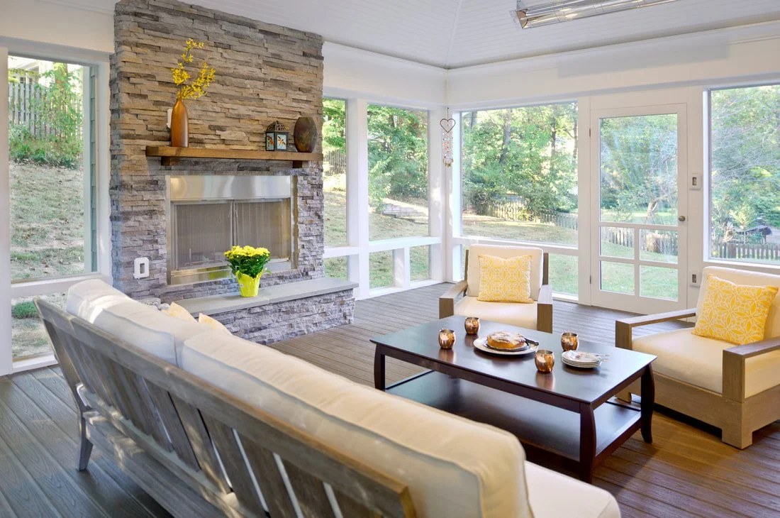 Sunroom with stone fireplace, wooden furniture, yellow cushions, and large windows overlooking a backyard.
