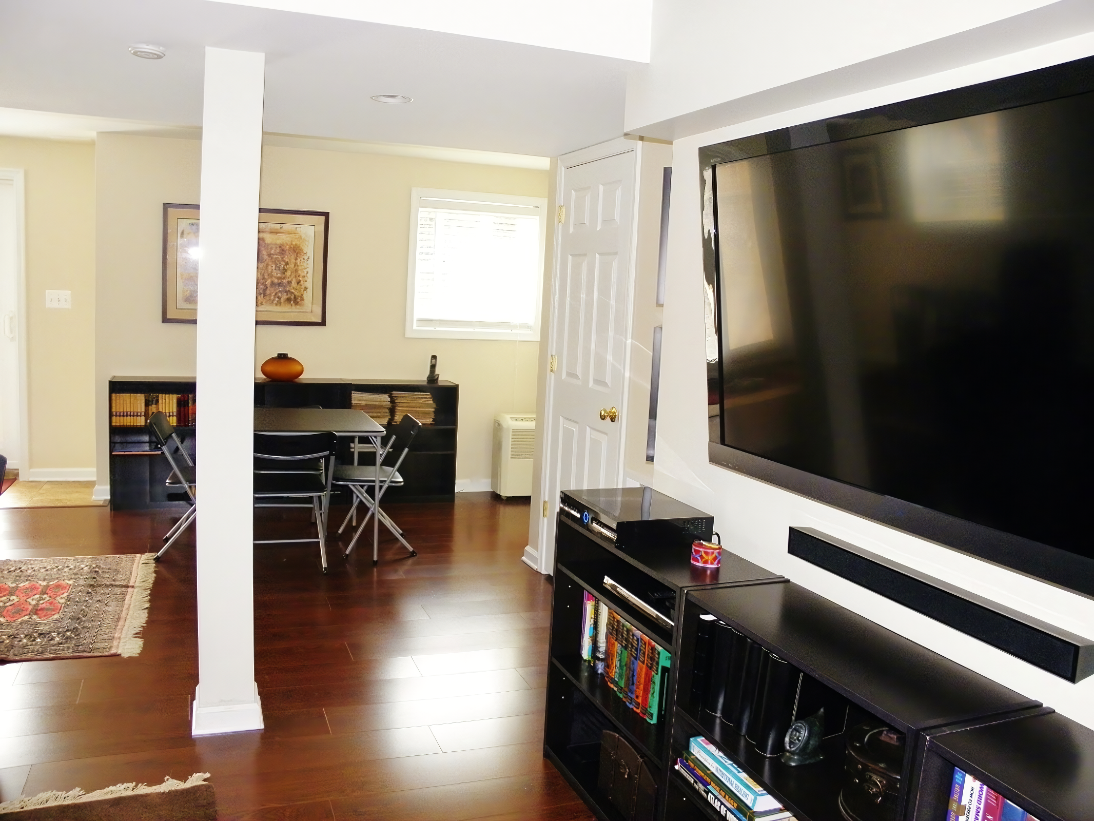 Living room with hardwood floors, a wall-mounted flat-screen TV, a black entertainment center, and a dining area with a black table and four black chairs. There's a window with blinds, artwork on the wall, and a small area rug.