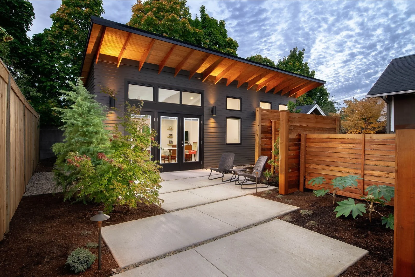 Modern backyard with concrete pathway, chairs, trees, and a black house with large windows and a wooden overhang.