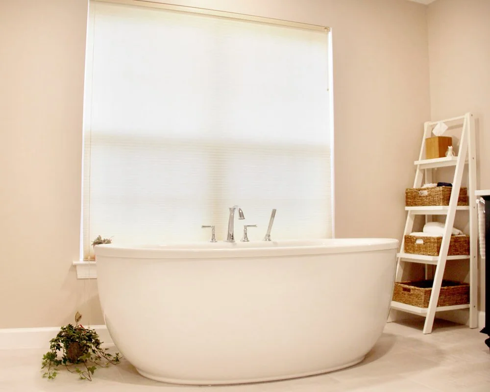 A modern bathroom with a white bathtub, a window with a shade, and a shelving unit with baskets and towels.