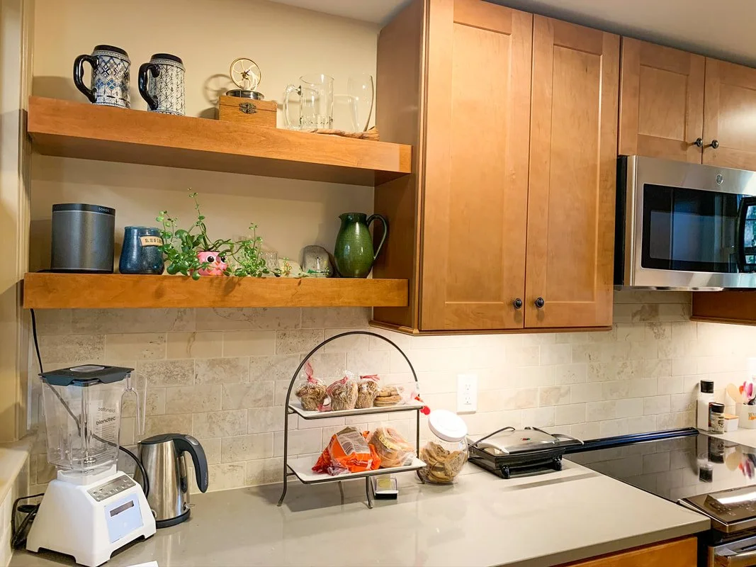Kitchen countertop with a blender, electric kettle, countertop organizer with snacks, and a grill. Open wooden shelves with decorative mugs, glasses, plants, and kitchen items. Wooden cabinets and a microwave oven.