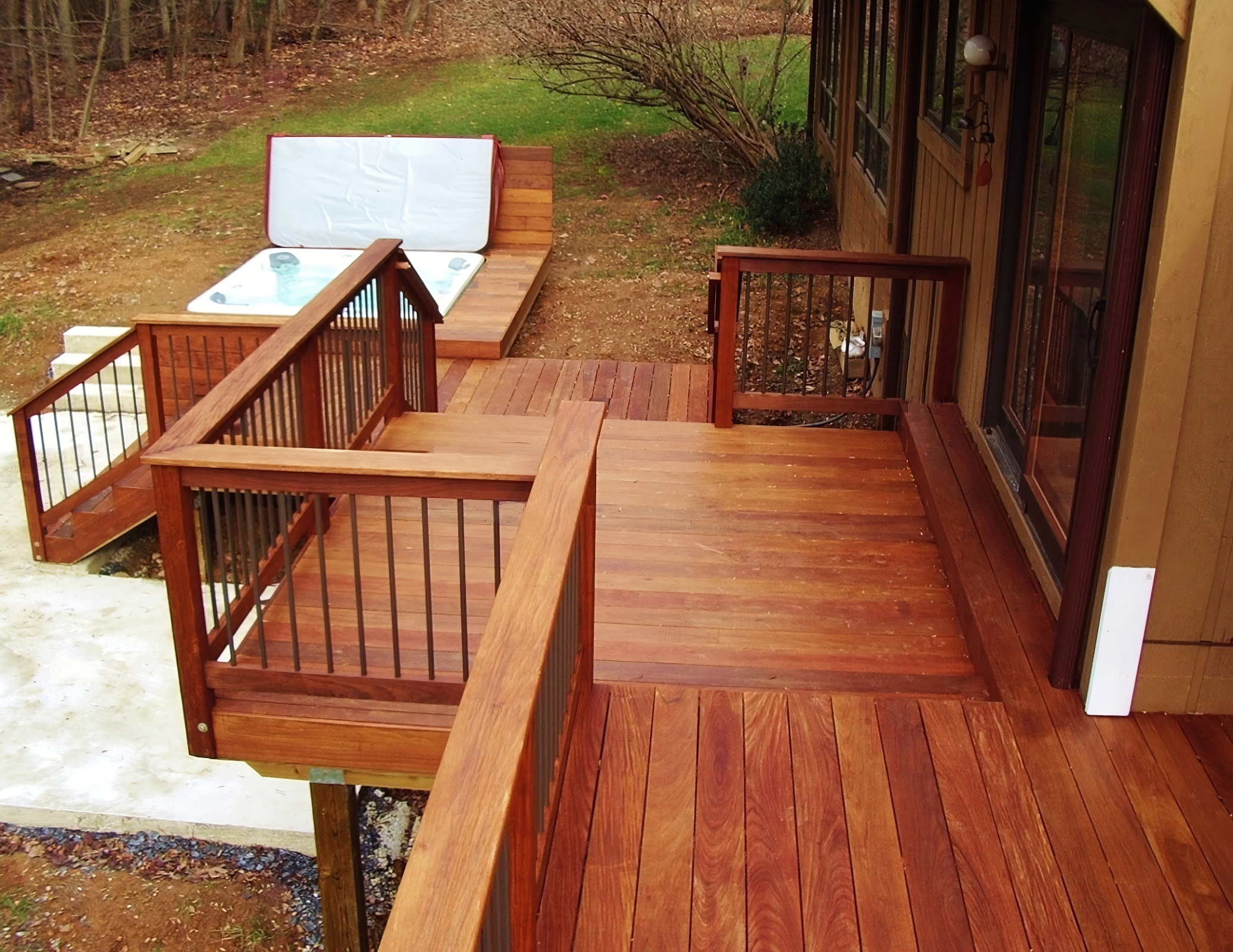 A wooden deck attached to a house with stairs leading down to a yard. The deck has a railing and a sliding glass door. The yard has a pile of snow and a wooded area in the background.