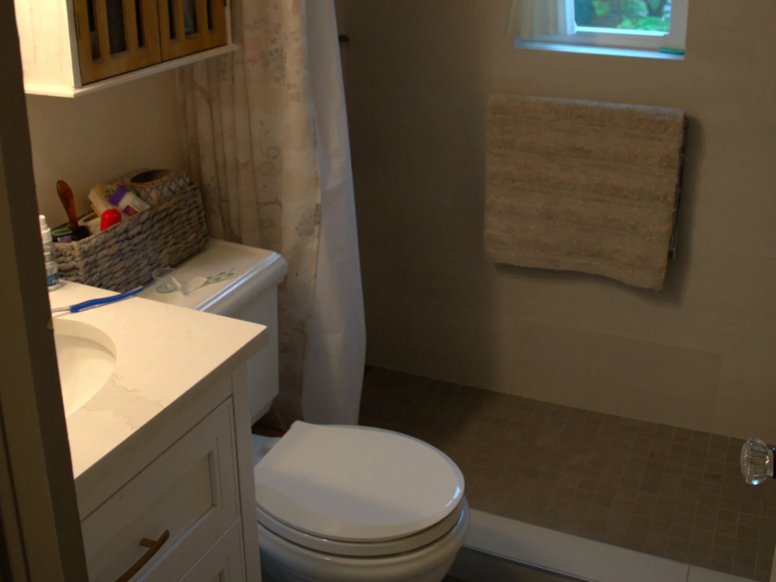 A small bathroom with a toilet, a vanity with a marble countertop, a wicker basket holding toiletries, a towel hanging on the wall, and a window above the toilet showing greenery outside.