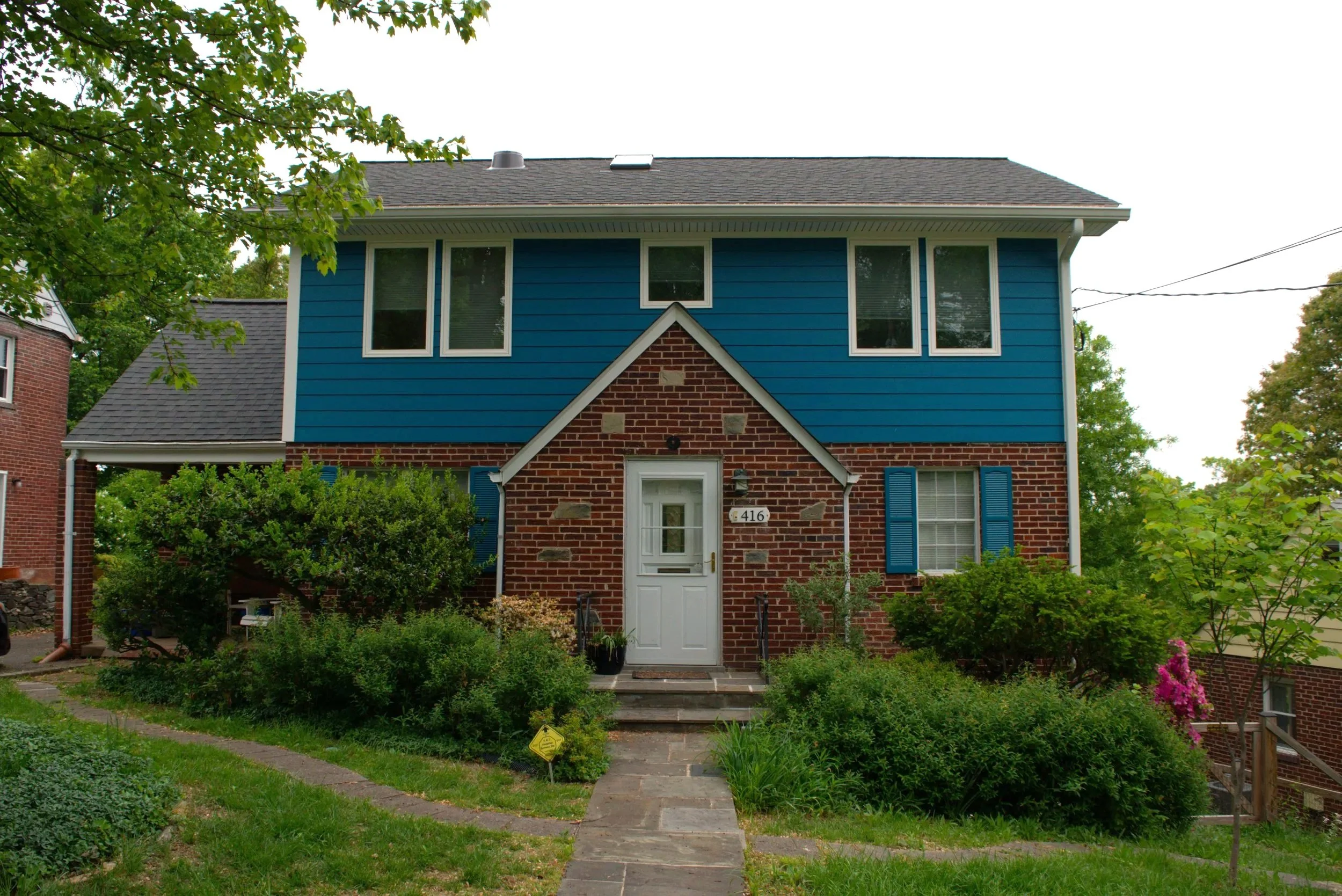 Front view of a house with a red brick lower level and blue upper level, white door and window shutters, surrounded by a green garden and trees.