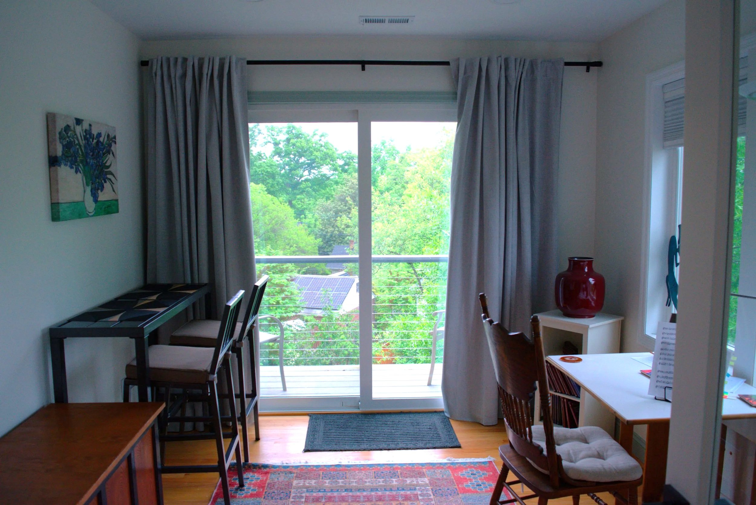 A living room with large windows and glass sliding doors looking out onto a green, leafy view. The room features a small table with a red vase, a desk, chairs, and a brightly patterned rug on hardwood floors. Curtains hang on either side of the slidi