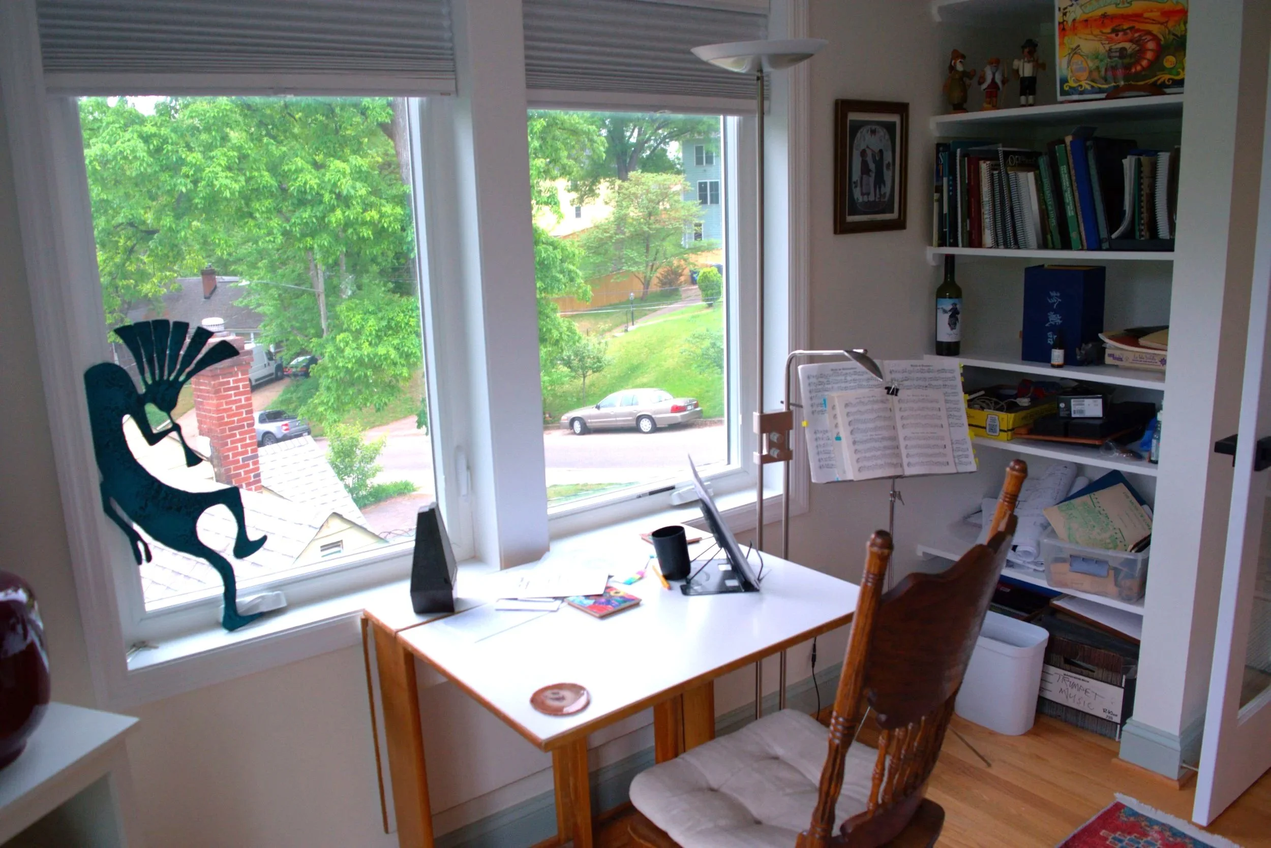 A cozy home office with a large window overlooking green trees and houses. There is a wooden desk with a music stand, sheet music, a computer monitor, and a speaker. A wooden chair with a white cushion is in front of the desk. To the right, a bookshe