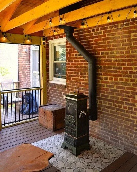 Indoor porch with brick walls, a wood stove, and string lights hanging from wooden beams.