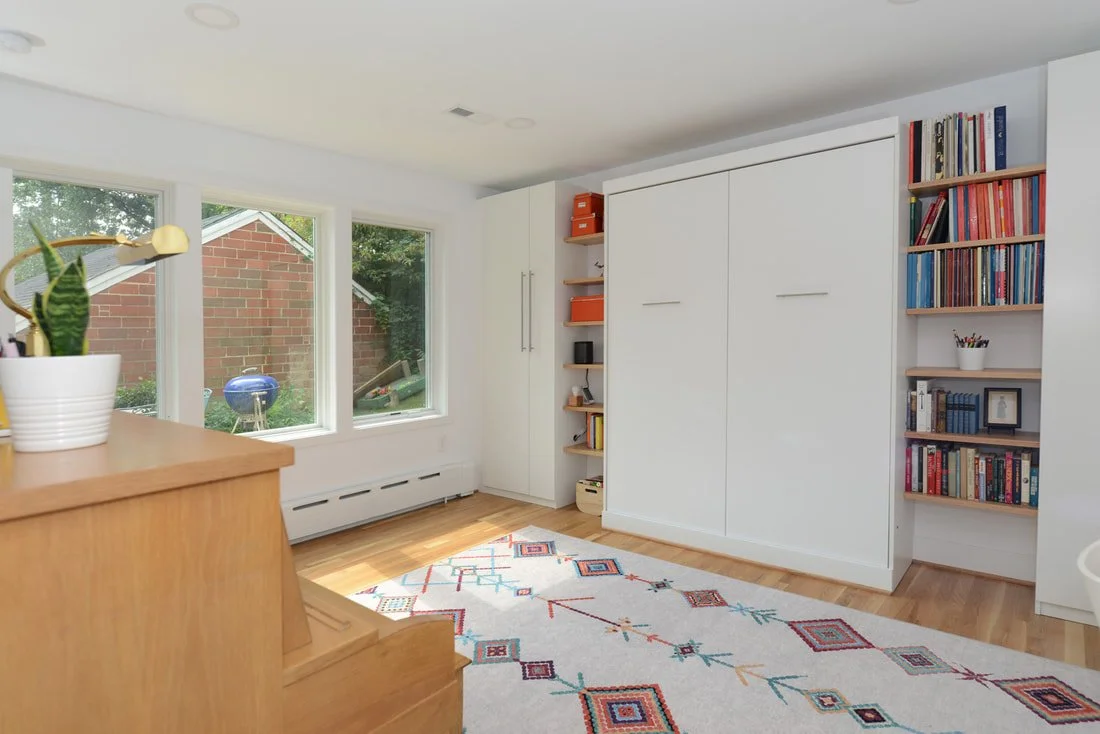 Bright living room with three large windows, wooden flooring, white walls, a white built-in closet, colorful patterned rug, and bookshelves filled with books on the right side.