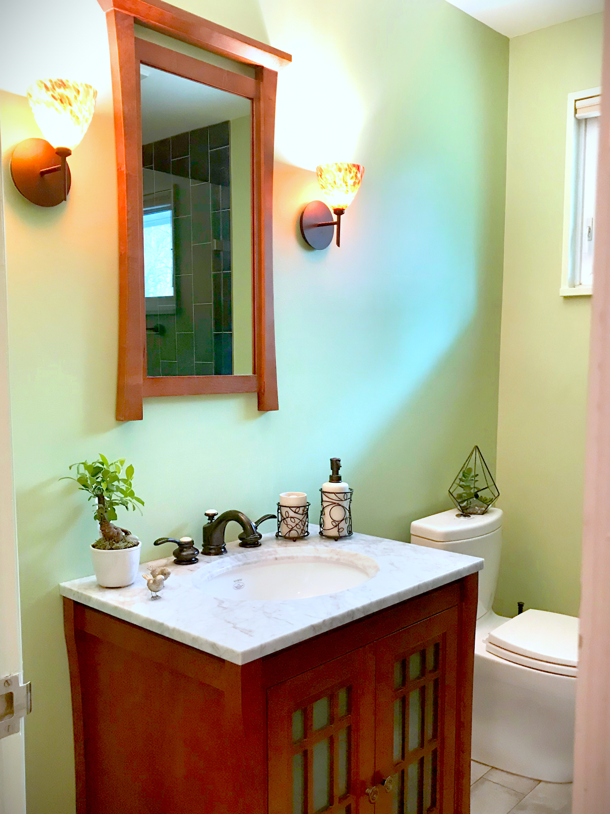 Bathroom with green walls, a wooden-framed mirror above a white marble vanity, a plant, soap dispenser, and decorative containers on the sink, and a white toilet with a geometric terrarium on top, illuminated by wall sconces.