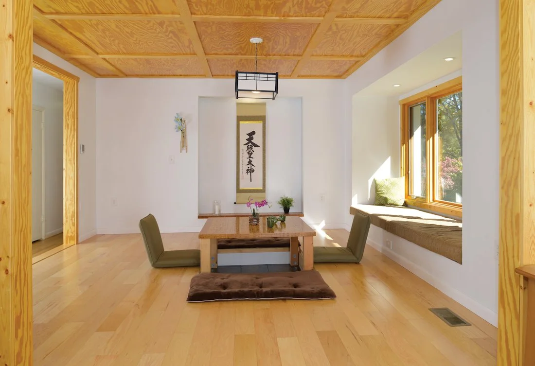 Minimalist Japanese-style dining area with a low wooden table, green floor cushions, a window seat with a pillow, and a decorative wall scroll.