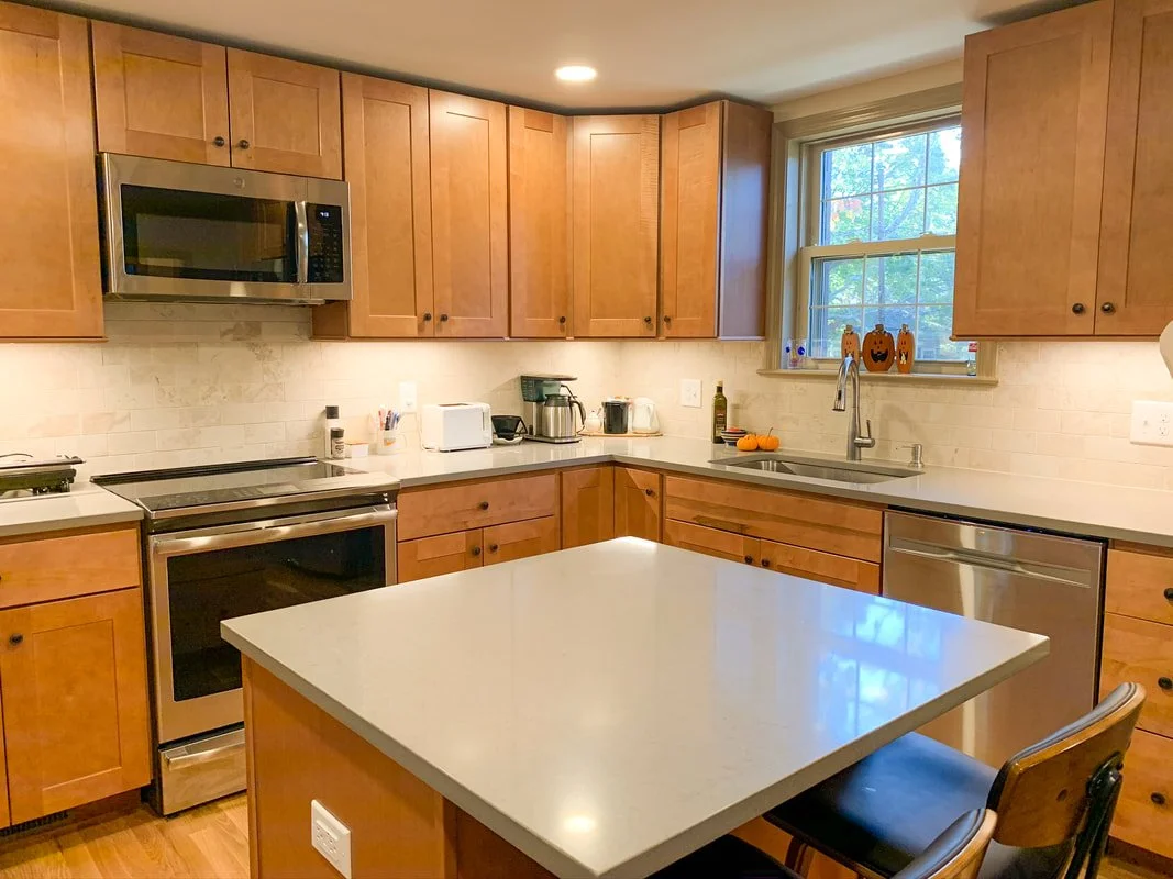 A kitchen with wooden cabinets, a white countertop island, and stainless steel appliances including a microwave, oven, and dishwasher. There's a window above the sink, some small decorative pumpkins, and various small appliances on the counter.