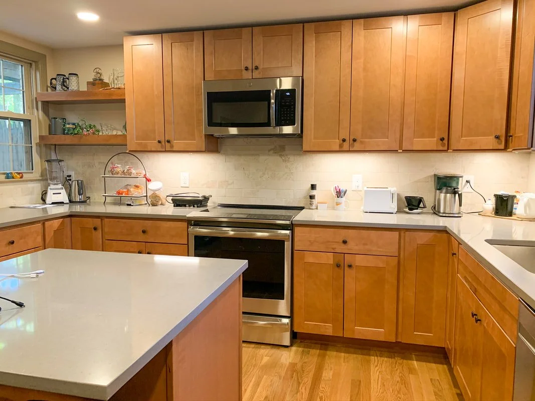 Kitchen with wooden cabinets, stainless steel microwave, oven, and stove, beige tile backsplash, white countertop, window, and various small appliances on the counter.