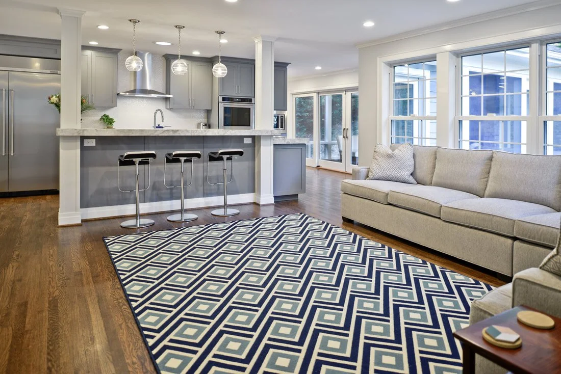 Open-concept living room and kitchen with gray cabinets, bar stools, a patterned rug, and large windows.