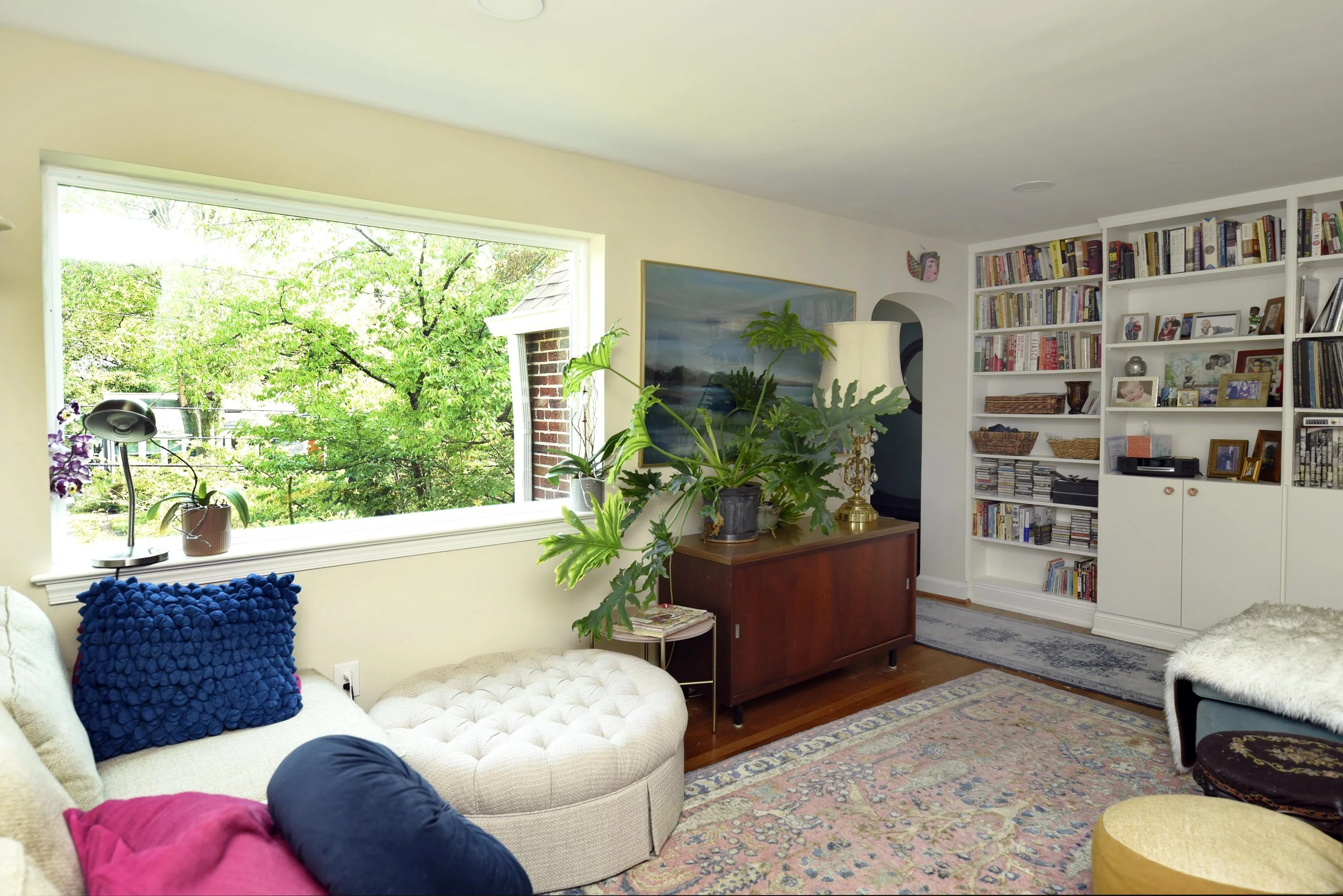 Living room with large window overlooking green trees, beige sofa with colorful pillows, wooden sideboard with plants and a lamp, built-in white bookshelf filled with books and decorative items, area rug, and a chair with a fur throw.