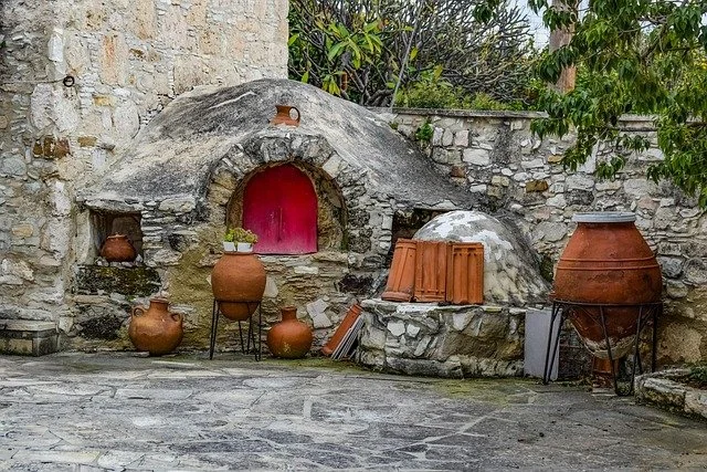A stone oven with a pink door, surrounded by pottery jars on patio stones, and greenery in background.