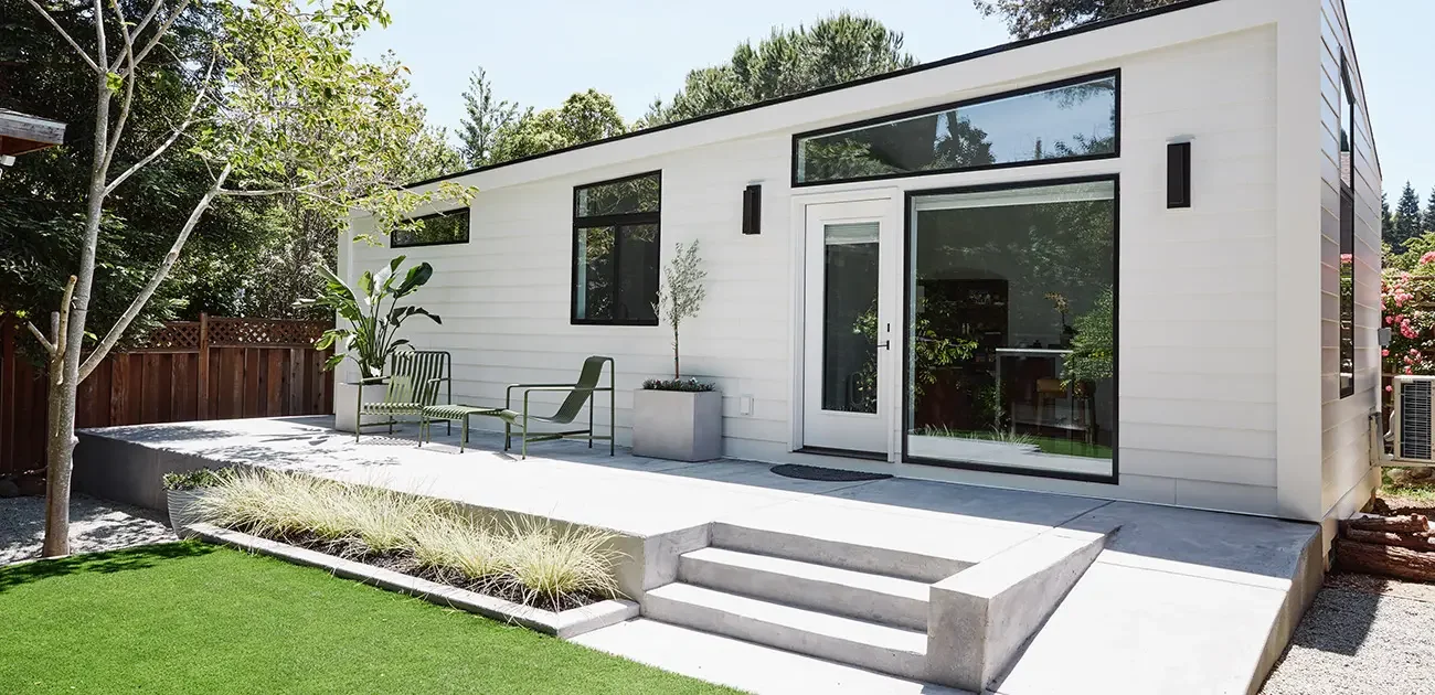 Modern white house with large glass sliding door, outdoor seating, potted plants, and landscaped yard with grass and trees.