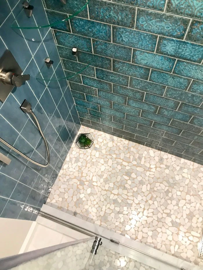 A shower with blue-green brick-patterned tiles on the wall, light-colored pebble-textured tiles on the floor, a small plant in a black pot in the corner, and glass shelves on the wall.