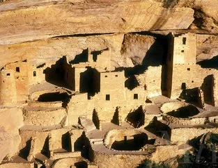Ancient cliff dwelling with multiple rectangular and circular rooms built into a sandstone cliff.