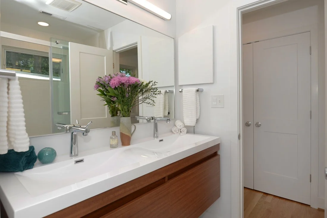 Modern bathroom with a double sink vanity, mirror, white walls, and a vase of pink flowers.
