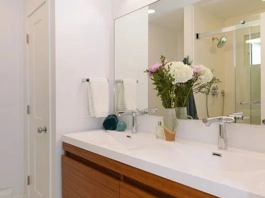 Bathroom with double sink, large mirror, vase of white and pink flowers, and a shower with glass door in the background.