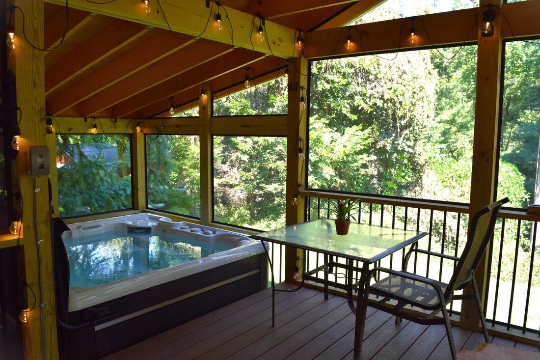 Screened porch with a hot tub, a small table with a potted plant, a chair, and string lights, overlooking a green forested area.