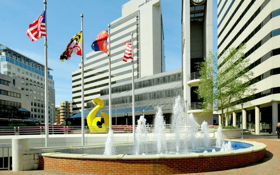 City square with a fountain, flagpoles with American and Maryland flags, modern tall office buildings, and a yellow sculpture.