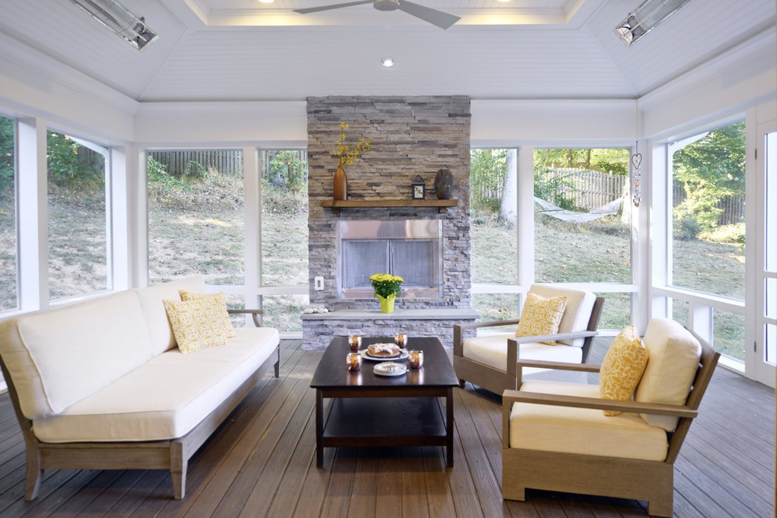Sunroom with beige sofas and armchairs, a black coffee table with snacks and drinks, a stone fireplace with decorative vases and a plant, large windows with a view of a grassy yard, a hammock outside, and a white ceiling fan.