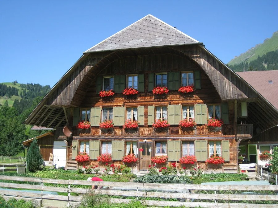 A traditional chalet-style wooden house with green shutters and red flower boxes, situated in a mountainous area with lush green hills and a clear blue sky.