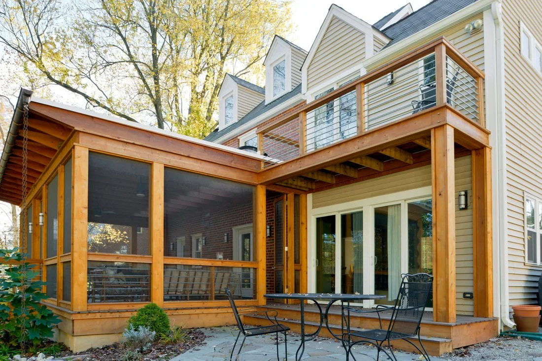 Back view of a house featuring a screened-in porch and an upper balcony with metal railing, with trees in the background.