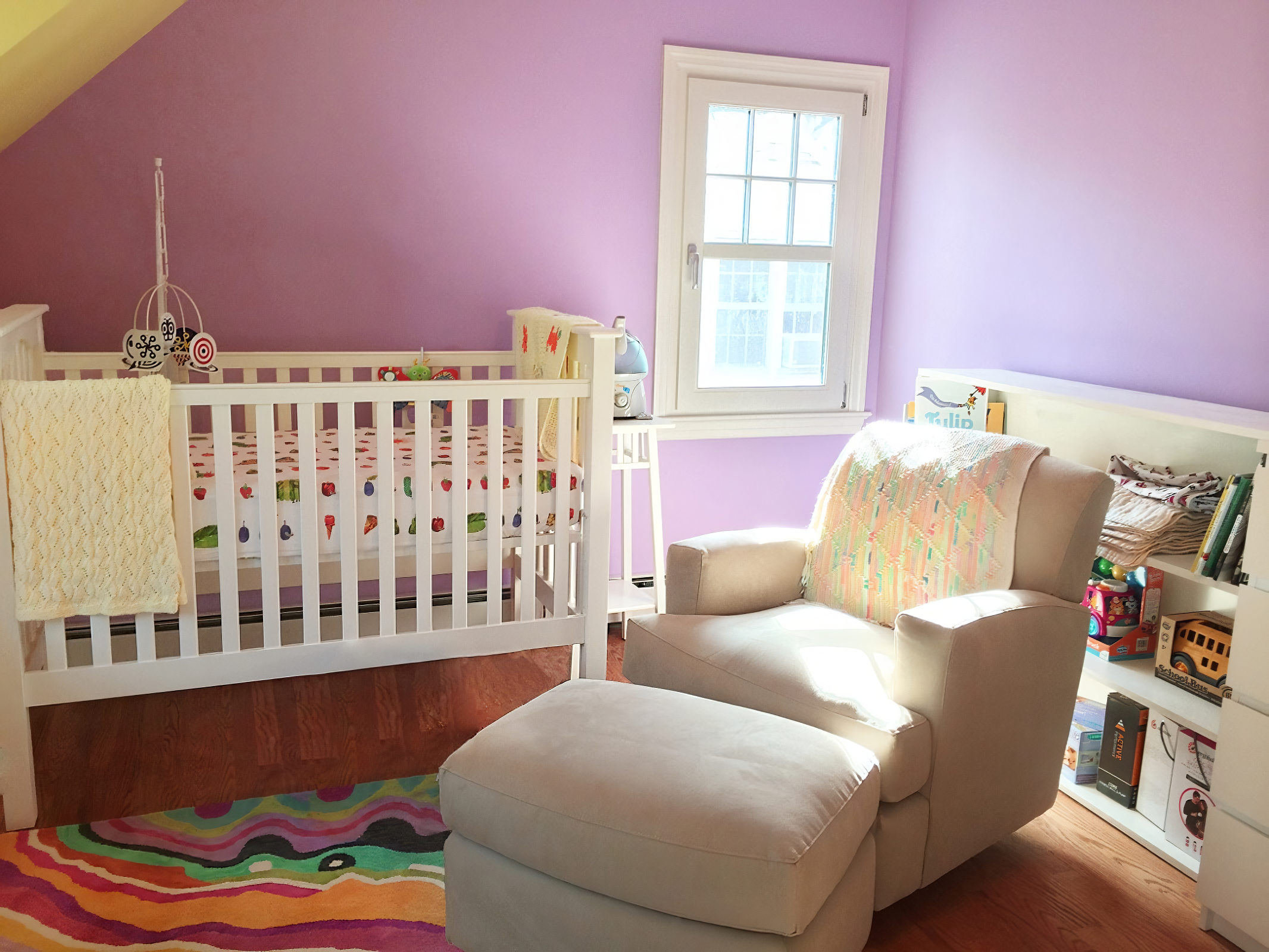 A cozy nursery with purple walls, a white crib with colorful bedding, a window allowing natural light, a beige armchair with an ottoman, and a white bookshelf with toys and books. A rainbow-colored rug is on the wooden floor.