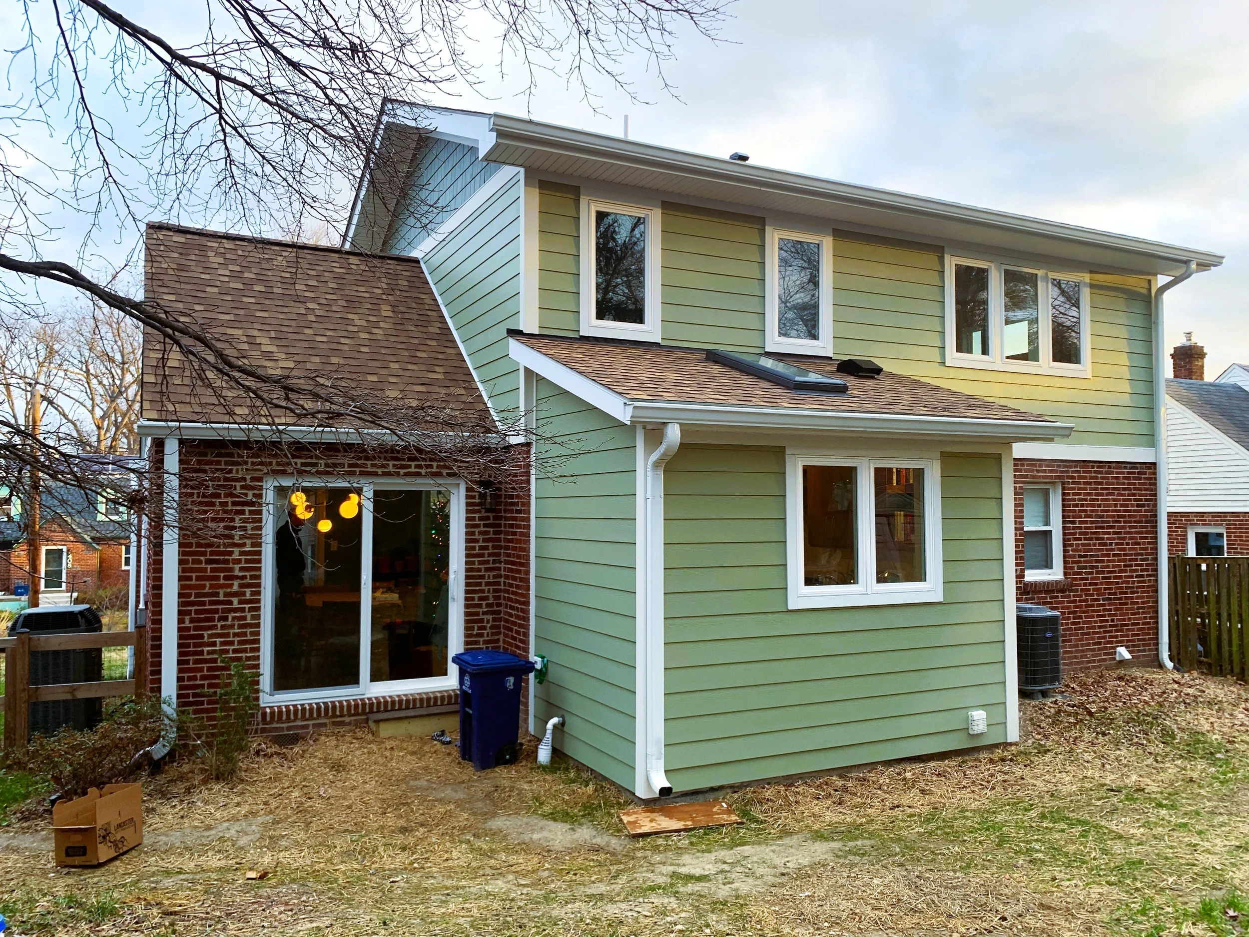 Back of a two-story house with green siding and brick accents, with several windows and a sliding glass door, a small patio area, and some yard debris.