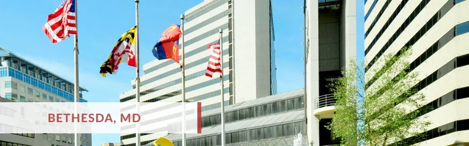 Multiple flags, including the American flag, flying in front of tall office buildings in Bethesda, Maryland, with a fountain and trees nearby.