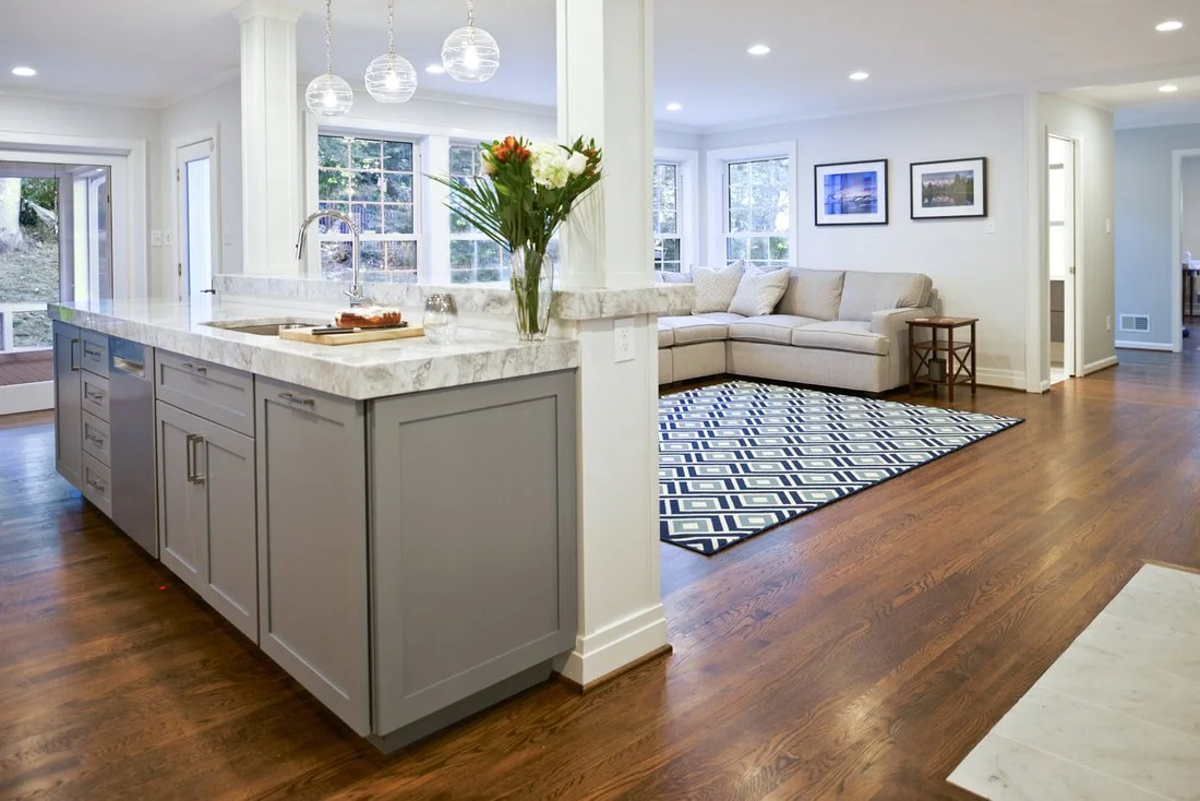 Open-concept living room and kitchen with white walls, wooden floors, a beige sectional sofa on a blue and white geometric rug, and a marble kitchen island with pendant lighting, fresh flowers, and modern decor.