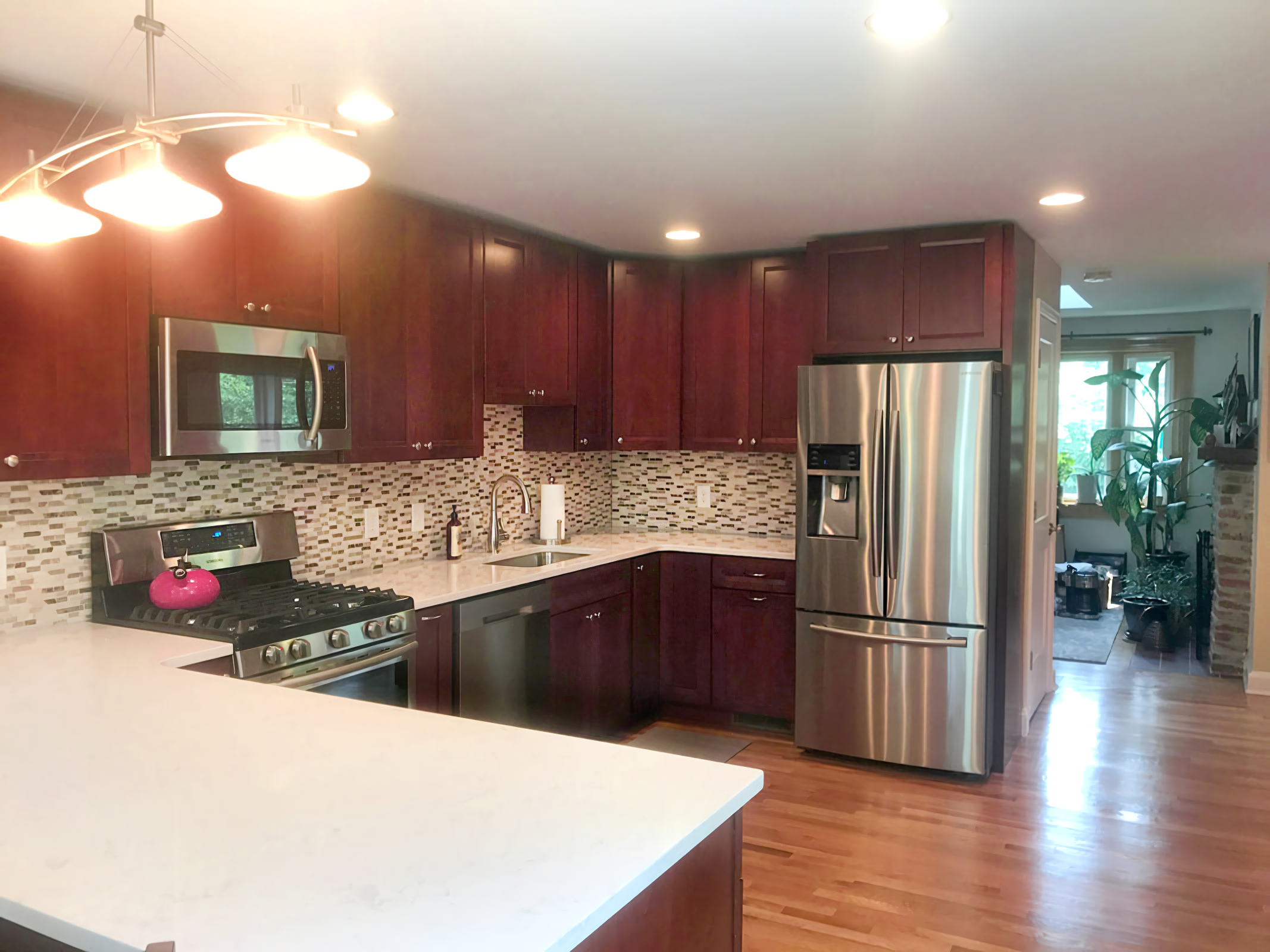Modern kitchen with dark wood cabinets, stainless steel appliances, mosaic tile backsplash, white countertops, hardwood floors, and a view into a sunlit living area with plants.