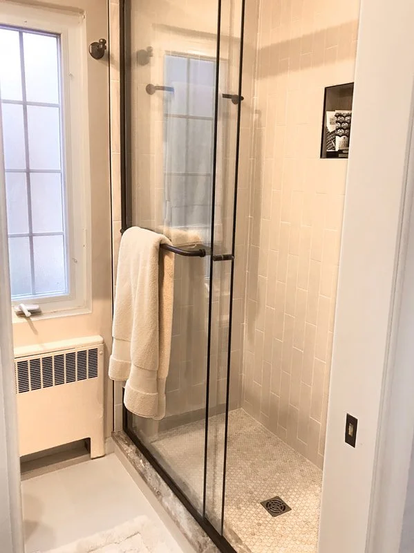 Bathroom with a glass shower enclosure, beige tiled walls, a window, a heater, and a towel hanging on the shower handle.