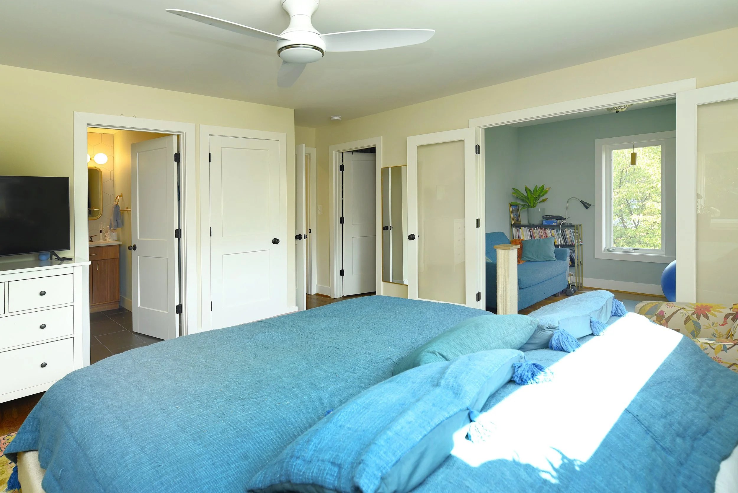 A bedroom with a blue bed and pillows, a ceiling fan, a white dresser with a TV, doors leading to other rooms, and a seating area with books, a lamp, and a window showing greenery outside.