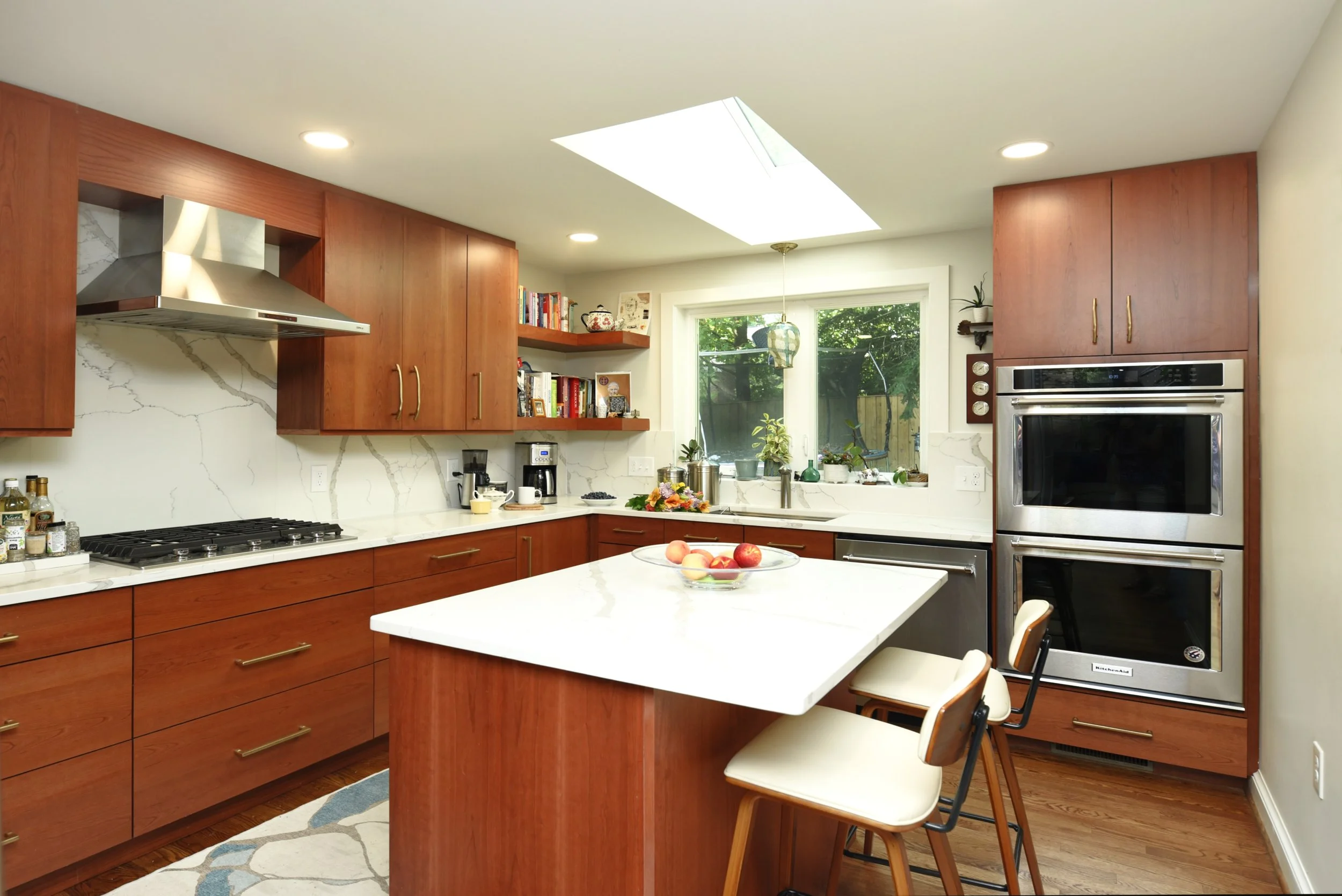 A modern kitchen with wooden cabinets, white marble countertops, and stainless steel appliances, including a double oven and a gas stove. There is a kitchen island with a bowl of apples, a window with plants outside, and open shelves with books and d